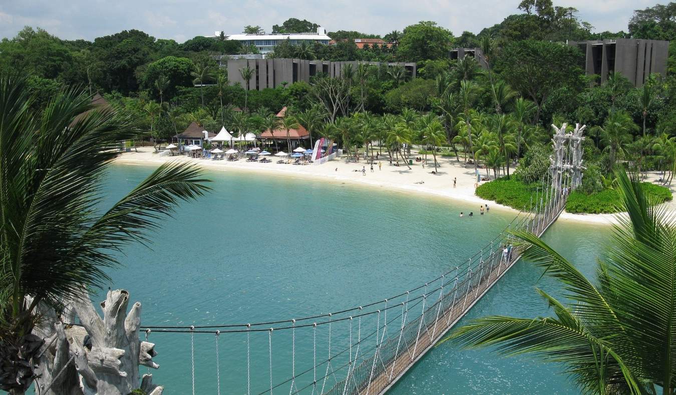 Un puente colgante peatonal que lleva a una playa de arena con palmeras y pequeñas cabañas en Sentosa, Singapur