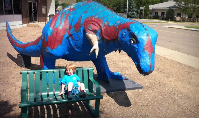 Un bebé llorando mientras está sentado en un banco cerca de una estatua azul de un dinosaurio mientras está de vacaciones