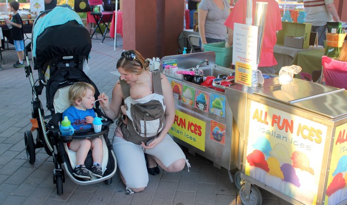 Una madre llevando a su bebé mientras da de comer a un niño pequeño sentado en un cochecito