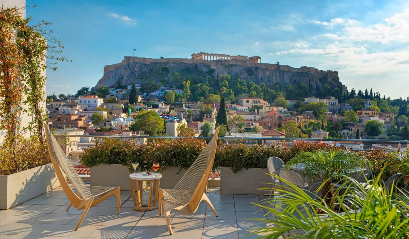 Butacas de madera frente a una mesa baja con copa de champán en la terraza de la azotea del hotel Plaka con vistas a la Acrópolis de Atenas, Grecia