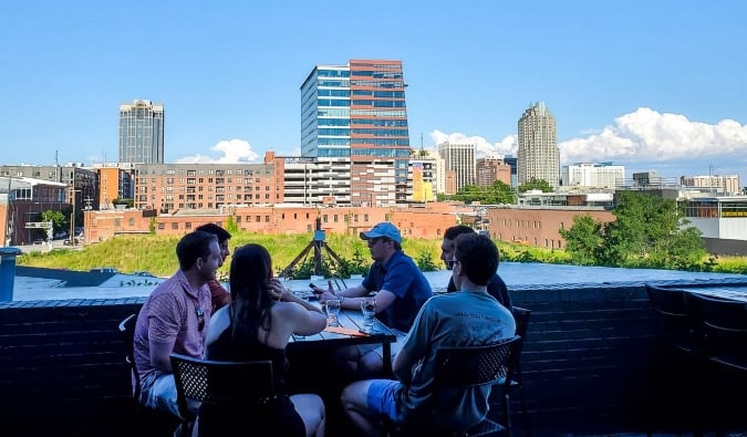 Un grupo de personas se sientan en una mesa en Wye Hill Kitchen & Brewing con el horizonte de Raleigh, Carolina del Norte al fondo