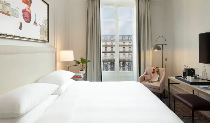 Una habitación de hotel parisino con una cama grande, pintura en la pared y una ventana abierta que muestra la arquitectura icónica de París al fondo