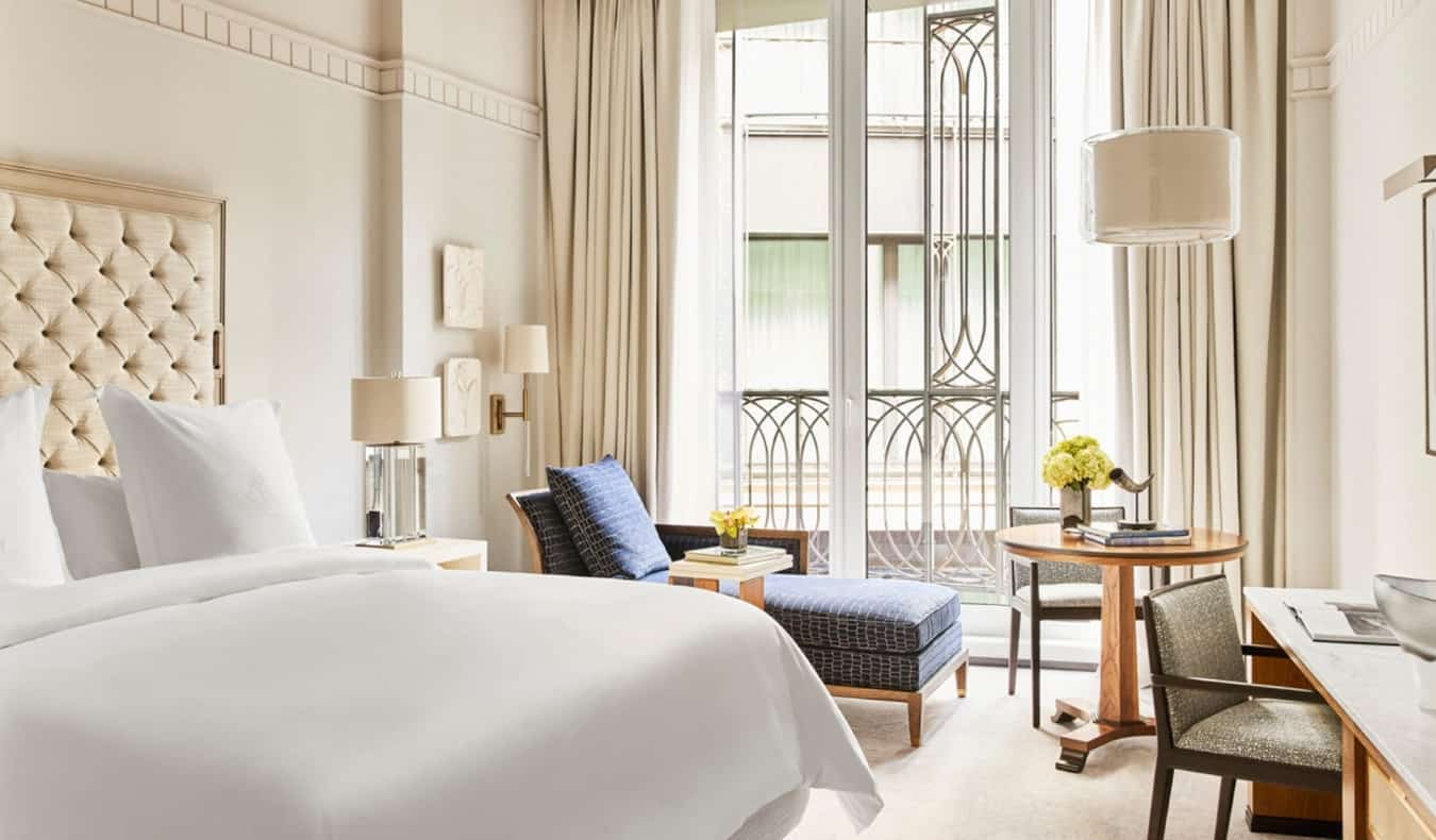 Una habitación de hotel de lujo de color crema con una cama grande, una chaise longue azul junto a la ventana abierta y un escritorio en el Four Seasons de Madrid, España