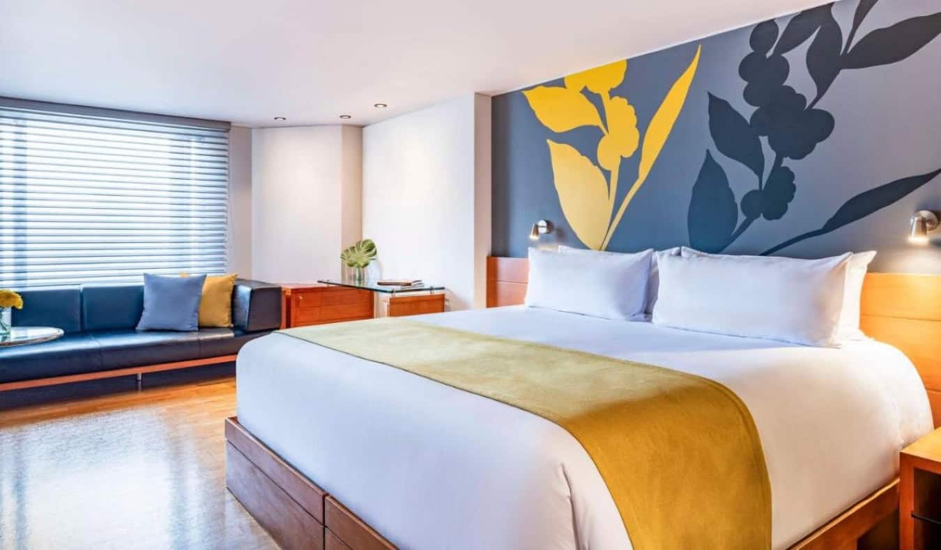 Cama grande en una habitación de invitados con una pared pintada con flores, suelos de madera dura y un gran banco con ventana en el hotel Avani Royal Zona T de Bogotá, Colombia