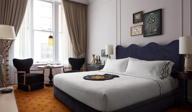 Una habitación de lujo con una cama grande, obras de arte enmarcadas en las paredes y una pequeña mesa redonda con sillas frente a una gran ventana enmarcada con cortinas en Maison Métier, un hotel de cinco estrellas de Nueva Orleans, Luisiana