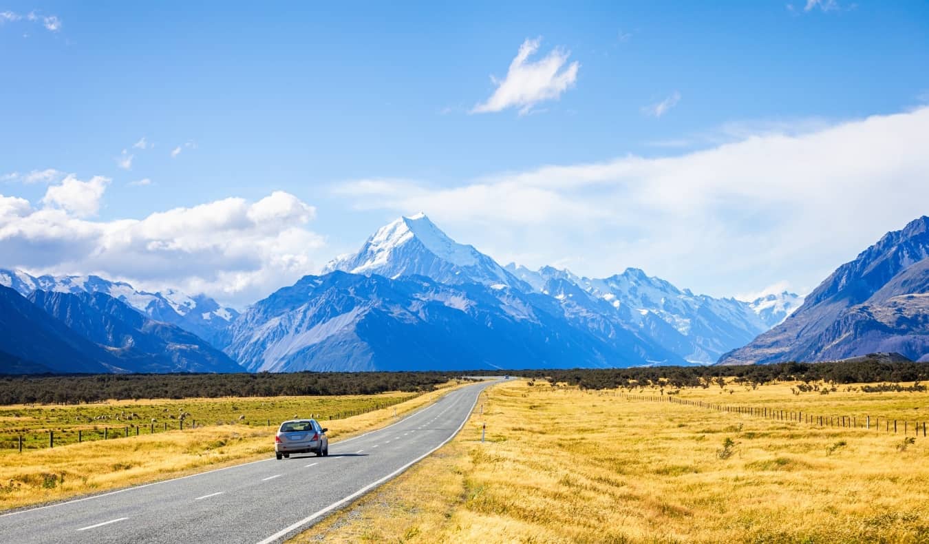 Coche conduciendo por una carretera con campos dorados a ambos lados, que conduce hacia montañas nevadas en la isla sur de Nueva Zelanda