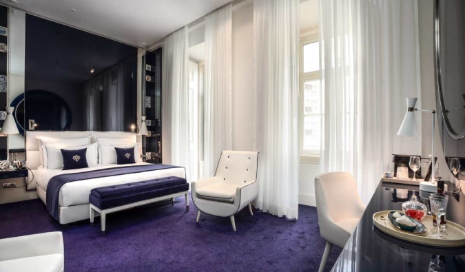 Una lujosa suite engalanada con alfombras de color azul intenso, un escritorio elegante, un sillón blanco, una cama grande y puertas francesas cubiertas con cortinas transparentes en Portugal Boutique Hotel de Lisboa, Portugal