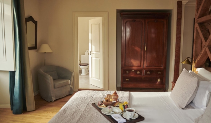 Una habitación de invitados con un gran armario de madera, suelos de parquet, una cama con una bandeja de comida para el desayuno y un pequeño sillón en la esquina del Hotel Alegría en Lisboa, Portugal